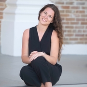 A smiling woman with long brown hair sits on outdoor stone steps in front of a brick building, wearing a black sleeveless outfit and black heels.