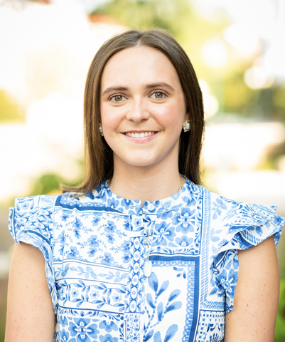Woman with dark brown hair wearing blue and white printed dress standing outside.