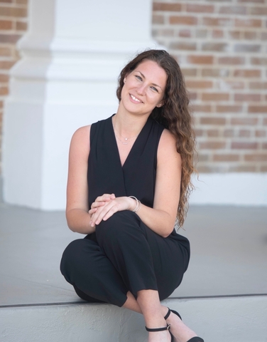 A smiling woman with long brown hair sits on outdoor stone steps in front of a brick building, wearing a black sleeveless outfit and black heels.