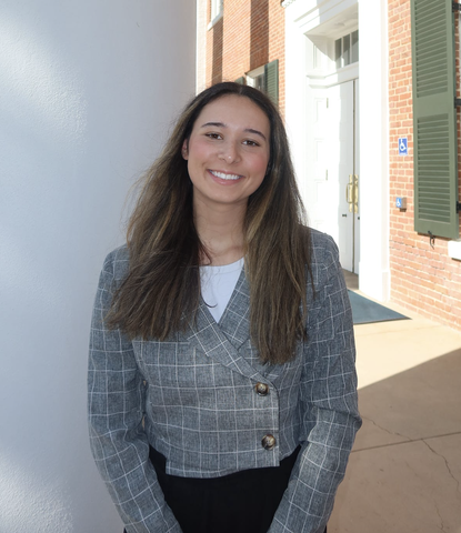 A smiling young woman in a blazer stands outside a building.