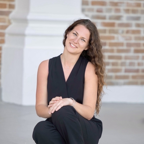 A smiling woman in a black dress sits on the ground in front of a brick wall.