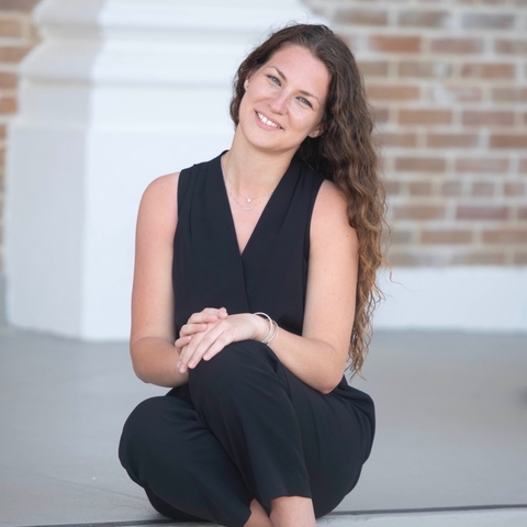 A smiling woman with long brown hair sits on outdoor stone steps in front of a brick building, wearing a black sleeveless outfit and black heels.