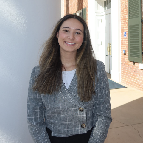 A smiling young woman in a blazer stands outside a building.