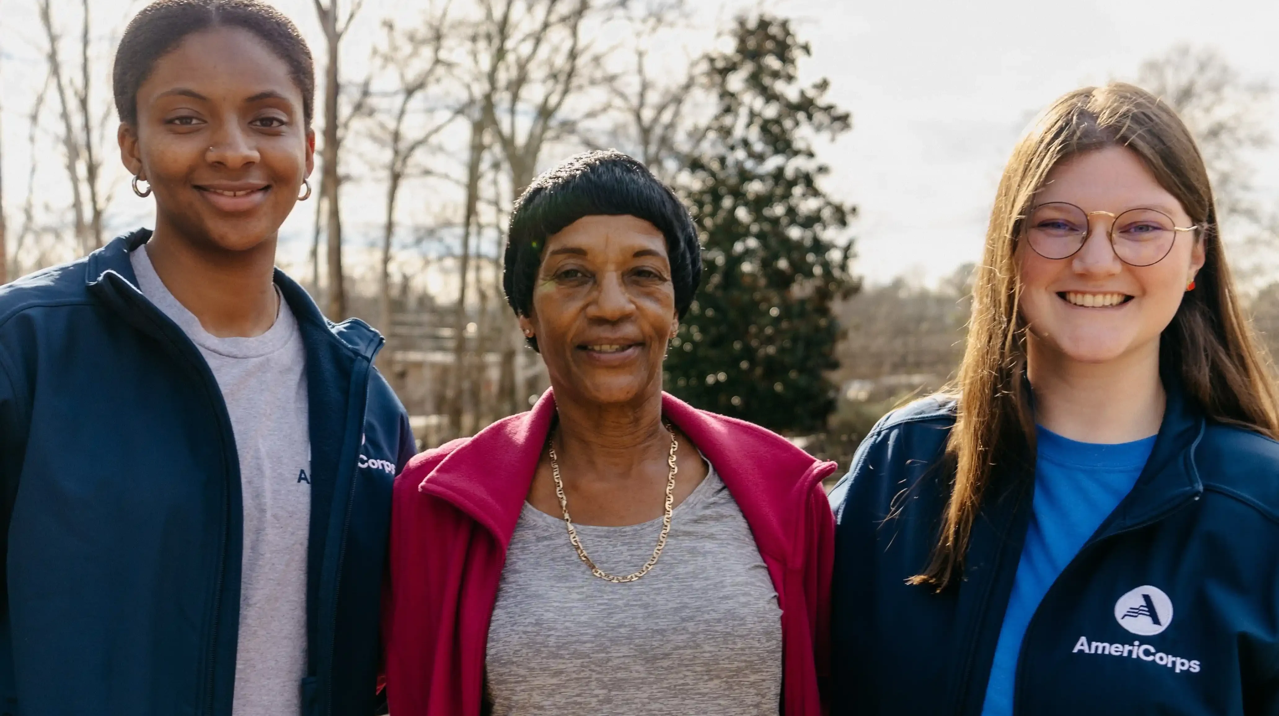 Three people standing outdoors smiling at camera. Person on right has on jacket with "AmeriCorps" written on it.